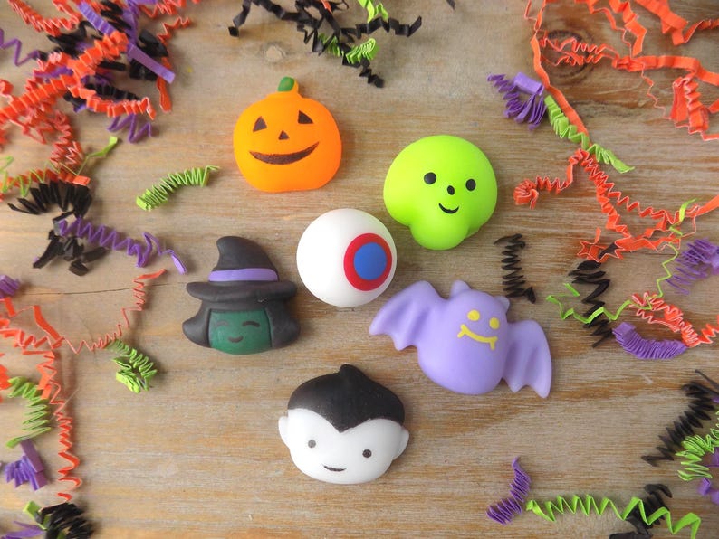 May include: A collection of six small Halloween-themed figurines including an orange jack-o'-lantern, a lime green skull, a white eyeball with a blue iris, a green-faced witch with a black hat, a purple bat, and a vampire with black hair.