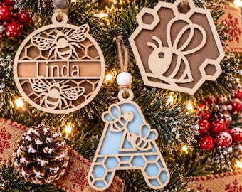 Pode incluir: Três ornamentos de madeira para pendurar com desenhos de abelhas e favos de mel. Um ornamento circular exibe o nome Linda, um ornamento hexagonal mostra uma abelha e um ornamento em forma de letra A apresenta duas abelhas, todos suspensos.