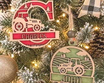 Peut inclure: Deux décorations de Noël en bois découpées au laser suspendues par de la ficelle. L'une est une lettre C rouge avec un tracteur et le nom Charles. L'autre est une décoration verte circulaire avec un tracteur dans un champ et le nom John.