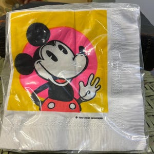 May include: White paper napkin with a yellow background and a pink circle featuring a cartoon image of Mickey Mouse. The text "Walt Disney Productions" is printed in black at the bottom of the napkin.