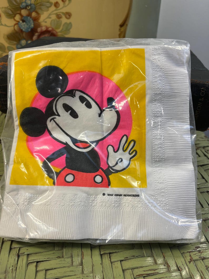 May include: White paper napkin with a yellow background and a pink circle featuring a cartoon image of Mickey Mouse. The text "Walt Disney Productions" is printed in black at the bottom of the napkin.