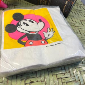 May include: A stack of white paper napkins with a yellow background and a cartoon image of Mickey Mouse waving. The image is surrounded by a pink circle. The text "WALT DISNEY PRODUCTIONS" is printed in black at the bottom of the napkin.