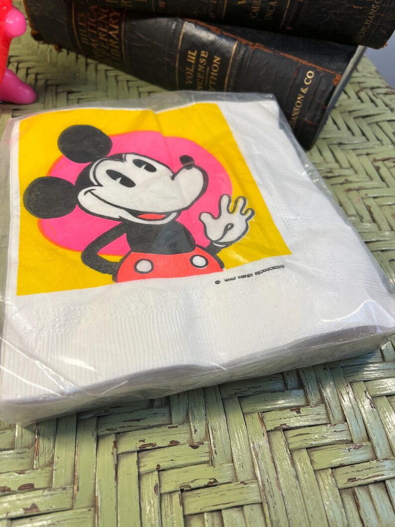 May include: A stack of white paper napkins with a yellow background and a cartoon image of Mickey Mouse waving. The image is surrounded by a pink circle. The text "WALT DISNEY PRODUCTIONS" is printed in black at the bottom of the napkin.
