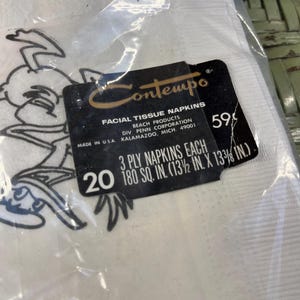 May include: A pack of 20 white facial tissue napkins featuring a cartoon character on the front. The pack is labelled "Contempo Facial Tissue Napkins" and "3 Ply Napkins Each 180 sq. in. (13 1/2 in. x 13 3/8 in.)".