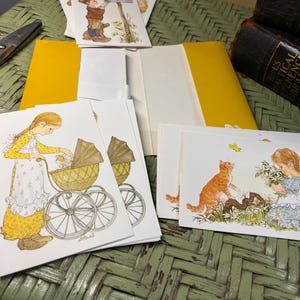 May include: A set of vintage note cards featuring illustrations of children. One card shows a girl in a yellow dress pushing a baby carriage. Another card shows a girl in a blue dress with a cat and a butterfly.
