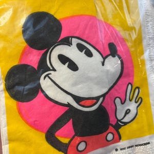 May include: A yellow and pink paper napkin featuring a cartoon image of Mickey Mouse. Mickey is wearing red shorts with white polka dots and is waving. The text "WALT DISNEY PRODUCTIONS" is in the bottom right corner.