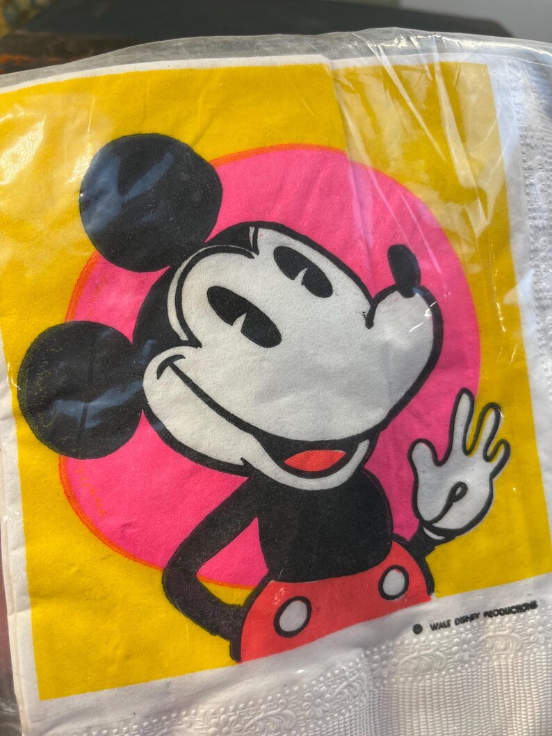 May include: A yellow and pink paper napkin featuring a cartoon image of Mickey Mouse. Mickey is wearing red shorts with white polka dots and is waving. The text "WALT DISNEY PRODUCTIONS" is in the bottom right corner.