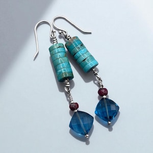 May include: A pair of dangle earrings featuring turquoise and blue gemstones. Each earring has a stack of turquoise disc beads, a small red bead, and a faceted blue square bead. The earrings have silver-colored ear wires.