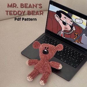 Può includere: Un orsacchiotto di peluche marrone lavorato a uncinetto seduto su un computer portatile. Lo schermo del computer portatile mostra un cartone animato di Mr. Bean che guida un'auto. Il testo sullo schermo del computer portatile dice "Mr. Bean's Teddy Bear Pdf Pattern".