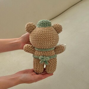 May include: A handmade, plush teddy bear in a sumo wrestler design. The bear is primarily tan with a green hat and belt. The bear is being held in someone's hands.