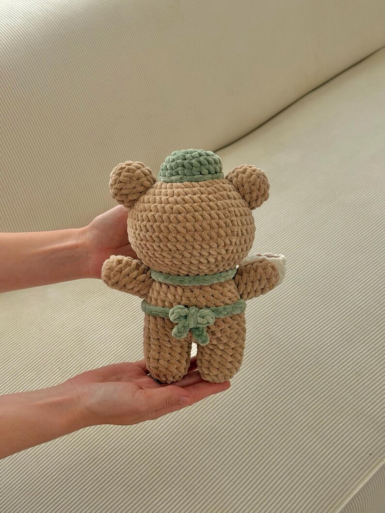 May include: A handmade, plush teddy bear in a sumo wrestler design. The bear is primarily tan with a green hat and belt. The bear is being held in someone's hands.