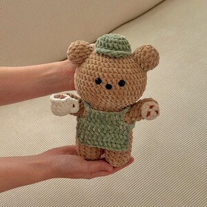May include: A handmade crochet bear doll in tan and green. The bear wears a green hat and overalls, and holds a white teacup. The bear has black eyes and small red accents on its paws. A charming, soft toy.