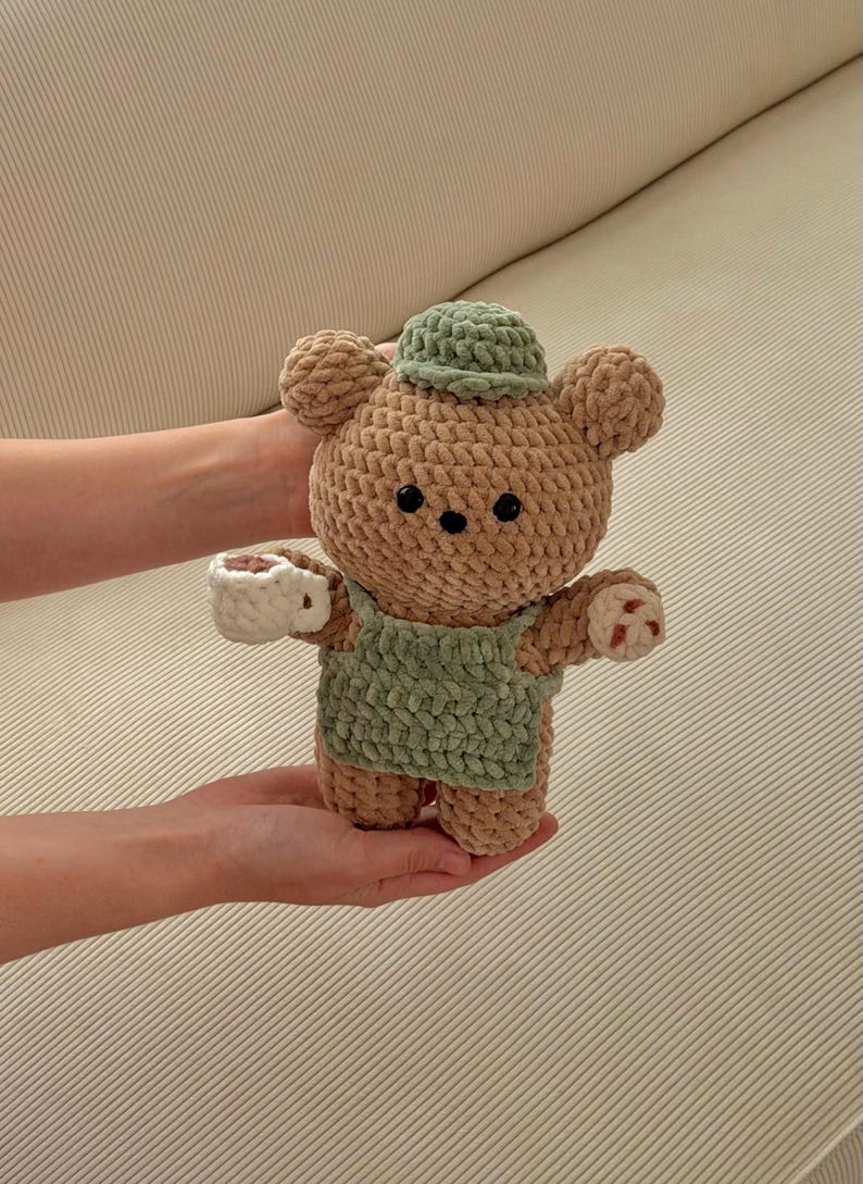 May include: A handmade crochet bear doll in tan and green. The bear wears a green hat and overalls, and holds a white teacup. The bear has black eyes and small red accents on its paws. A charming, soft toy.