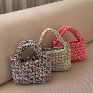 May include: Three small, handmade crochet bags in shades of pink, gray, and yellow. Each bag has a sturdy handle and a textured, woven appearance. The bags are displayed on a neutral-toned surface.