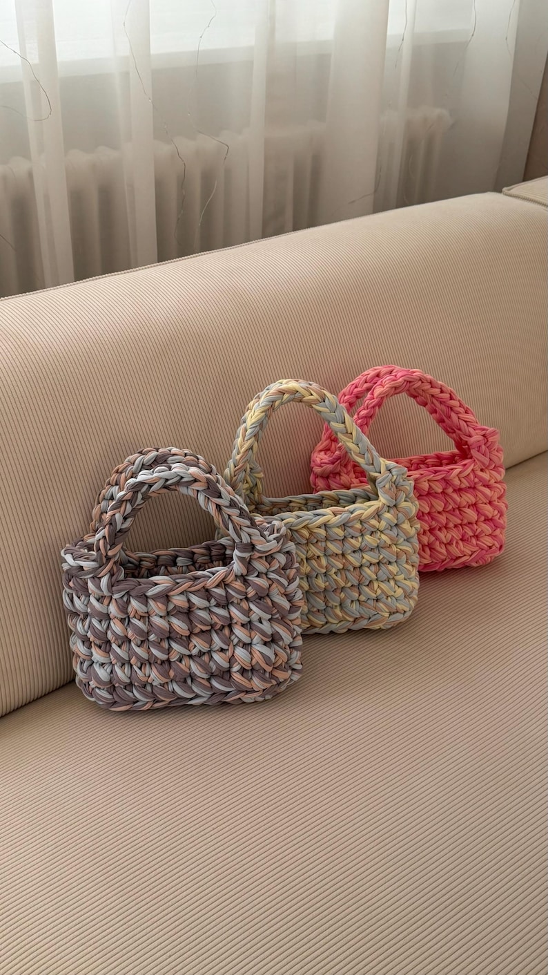 May include: Three small, handmade crochet bags in shades of pink, gray, and yellow. Each bag has a sturdy handle and a textured, woven appearance. The bags are displayed on a neutral-toned surface.