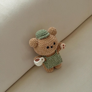 May include: A crocheted bear doll in tan and green, wearing a green apron and hat, holding a white cup with a brown liquid. The bear has black eyes and small red accents on its paws. The doll is handmade.