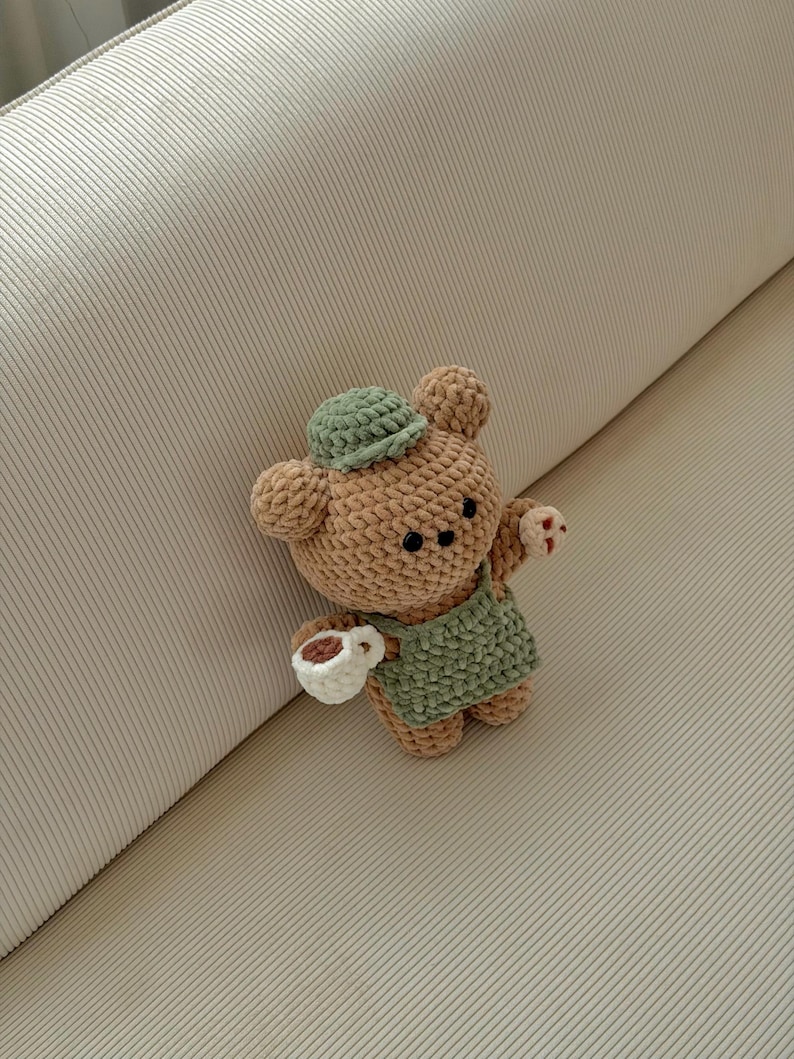 May include: A crocheted bear doll in tan and green, wearing a green apron and hat, holding a white cup with a brown liquid. The bear has black eyes and small red accents on its paws. The doll is handmade.