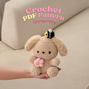 Può includere: Un cucciolo beige all'uncinetto con orecchie flosce, un fiore rosa e un'ape sulla testa. Il testo "Crochet PDF Pattern Spring puppy" è visualizzato in rosa e giallo. Il cucciolo è tenuto in una mano.