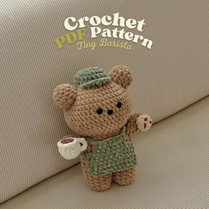 May include: A crocheted teddy bear wearing a green apron and hat, holding a white cup with a brown liquid. The bear is brown with black button eyes. The text "Crochet PDF Pattern Tiny Barista" is in the upper part of the image.