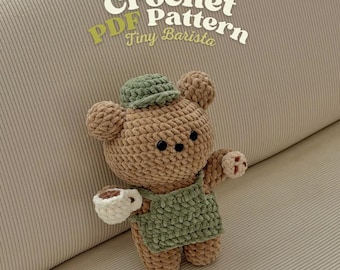 May include: A crocheted teddy bear wearing a green apron and hat, holding a white cup with a brown liquid. The bear is brown with black button eyes. The text "Crochet PDF Pattern Tiny Barista" is in the upper part of the image.