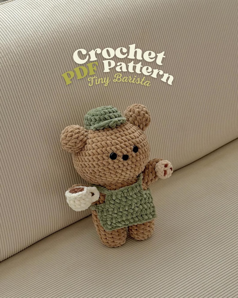 May include: A crocheted teddy bear wearing a green apron and hat, holding a white cup with a brown liquid. The bear is brown with black button eyes. The text "Crochet PDF Pattern Tiny Barista" is in the upper part of the image.