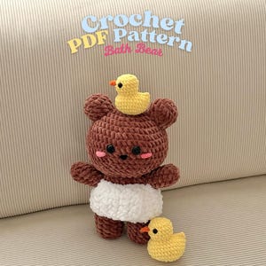 May include: A crocheted brown bear with a white nappy and a yellow duck on its head and another one beside it. The bear has black eyes and pink cheeks. The text "Crochet PDF Pattern Bath Bear" is above the bear.