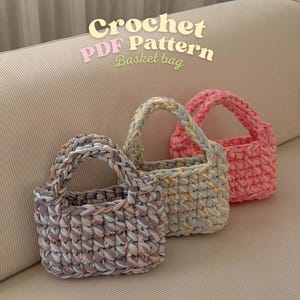 Könnte beinhalten: Drei gehäkelte Korbtaschen in Rosa, Grau und Gelb. Jede Tasche hat einen geflochtenen Henkel und ein strukturiertes Stichmuster. Der Text "Crochet PDF Pattern Basket bag" ist über den Taschen zu sehen.