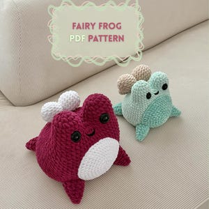 Può includere: Due giocattoli a forma di rana all'uncinetto. Uno è di colore magenta scuro con dettagli bianchi, e l'altro è azzurro con dettagli beige. Il testo "FAIRY FROG PDF PATTERN" è visualizzato in una cornice decorativa.
