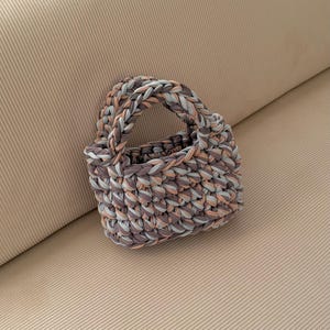 May include: A small, hand-knitted tote bag in shades of gray, peach, and light blue. The bag features a braided handle and a textured, woven appearance. The bag is sitting on a beige surface.