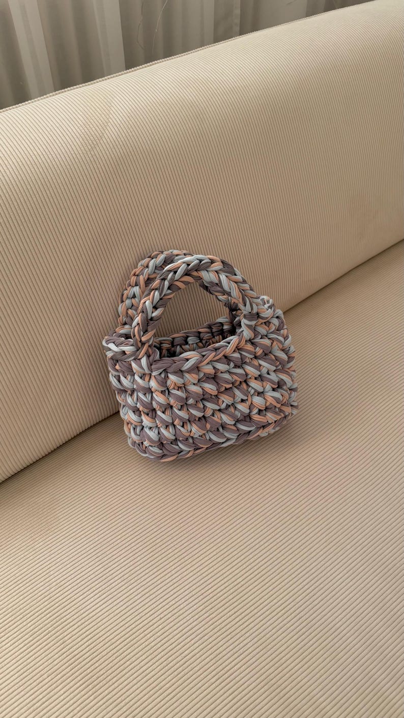 May include: A small, hand-knitted tote bag in shades of gray, peach, and light blue. The bag features a braided handle and a textured, woven appearance. The bag is sitting on a beige surface.