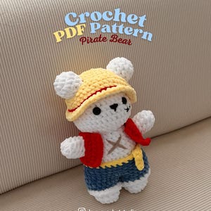 K&ouml;nnte beinhalten: Ein geh&auml;kelter wei&szlig;er B&auml;r mit gelbem Hut und rotem Band, roter Weste und blauen Shorts. Der B&auml;r hat einen gelben G&uuml;rtel und ein Kreuz auf der Brust. Der Text auf dem Bild lautet "Crochet PDF Pattern Pirate Bear."