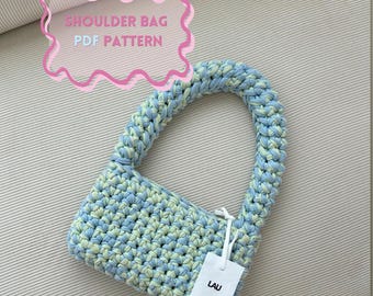 Op de afbeelding: Een handgemaakte schoudertas met een geweven textuur in blauw en groen. De tas heeft een gebogen handvat en een kleine rechthoekige body. De tekst "SHOULDER BAG PDF PATTERN" staat in een roze wolk. Een wit label leest "LAU".