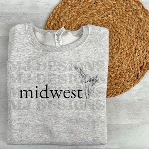 May include: A light gray sweatshirt with the word "midwest" in black script, accompanied by a black and white wheat stalk graphic. The sweatshirt is folded on a round, woven, brown placemat.