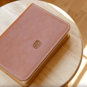 May include: A brown leather-bound journal with a stitched edge and a small embossed logo on the front cover. The journal is sitting on a wooden surface.