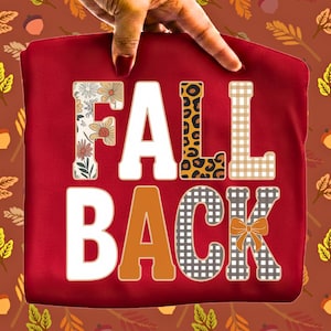 May include: A dark red sweatshirt featuring the text FALL BACK in large block letters. The letters are decorated with various patterns including floral prints, leopard spots, tan gingham, solid orange, and a gray plaid with a small orange bow.