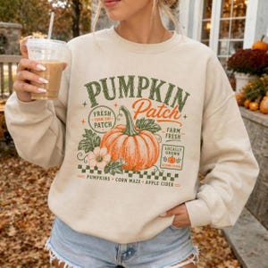 May include: A beige crewneck sweatshirt featuring a graphic of a large orange pumpkin with green leaves. The text reads PUMPKIN Patch, FRESH FROM THE PATCH, FARM FRESH, LOCALLY GROWN, and PUMPKINS CORN MAZE APPLE CIDER in green and orange lettering.