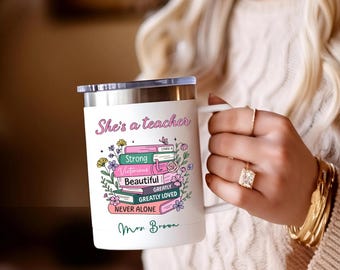 May include: White travel mug with a silver lid and handle. The mug features the text "She's a teacher" above a stack of books with the words "Strong, Victorious, Beautiful, Greatly Loved, Never Alone". Floral and butterfly accents complete the design.