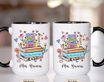 May include: White ceramic mug with a black handle and rim. The mug features a colorful design with books, pencils, flowers, and the text "Helping Little Minds Grow" and "#teacherlife". The name "Mrs. Brown" is also printed on the mug.