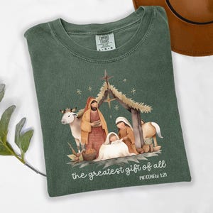 May include: Green Comfort Colors t-shirt featuring a watercolor illustration of the Nativity scene. The design includes Mary, Joseph, baby Jesus, and animals. The text "the greatest gift of all" and "MATTHEW 1:21" are printed below the image.
