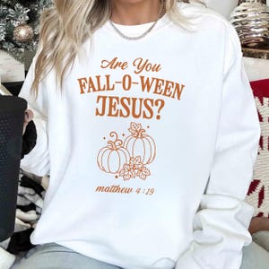 May include: White sweatshirt with the text "Are You Fall-O-Ween Jesus?" in orange. Below the text is an illustration of two pumpkins and leaves. The Bible verse Matthew 4:19 is printed below the illustration.