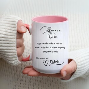 May include: White ceramic mug with a pink interior and handle. The mug features the text "Difference Maker" in a script font, along with a definition and the name "Michelle". The mug is held by a person wearing a white sweater.