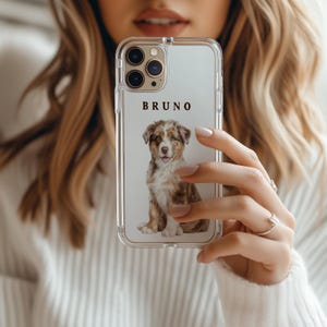May include: A clear smartphone case featuring a printed illustration of a brown and white Australian Shepherd puppy sitting down. The name BRUNO is printed in dark brown capital letters above the puppy graphic on the transparent case.
