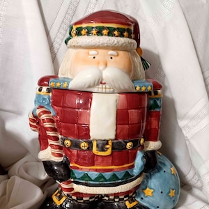 May include: A ceramic Santa Claus cookie jar with a red and green plaid pattern, a white beard, and a blue bag with gold stars.