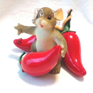 May include: A whimsical figurine of a mouse with a light brown body and cream-colored belly, holding three glossy red chili peppers. The mouse has clear plastic ears and is posed with one arm outstretched.