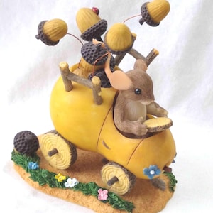 May include: A yellow acorn-shaped car with a brown mouse driving. The car has wooden wheels and is decorated with acorns and flowers. The mouse is holding a steering wheel.