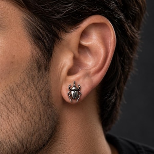 May include: A silver-toned metal stud earring shaped like a beetle. The insect design features a rounded body, a small head, and six distinct legs extending from the sides. The surface has a polished, reflective finish with dark recessed details.
