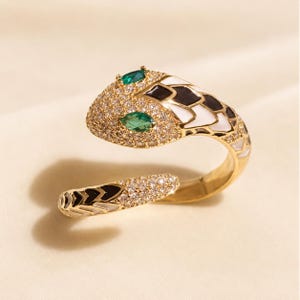 May include: A gold-toned snake ring featuring a wrap-around design. The snake head is encrusted with clear pavé stones, two marquise-cut green stones for eyes, and alternating black and white enamel scales along the body and tail section.