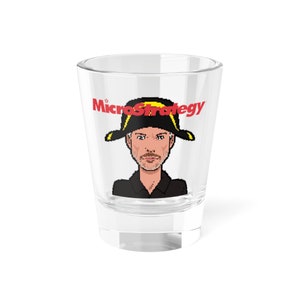 May include: A clear glass shot glass with a pixelated image of a man wearing a black hat with a yellow brim and a black shirt. The text "MicroStrategy" is printed in red on the glass.