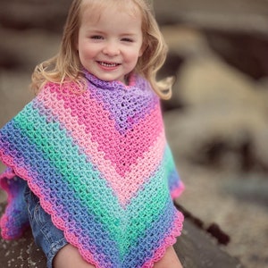 May include: A crochet poncho featuring a vibrant gradient of colours including pink, purple, blue, and green. The garment has a V-shaped hemline with a scalloped edge and a textured stitch pattern that creates a chevron design across the fabric.