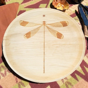 May include: Round, light-colored wooden plate with a dragonfly design. The dragonfly has brown wings and body, with a star-like design above it. The plate is likely used for serving food.